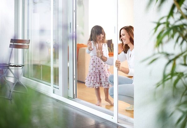 Mutter und Tochter lächeln hinter Insektenschutz-Schiebetür im modernen Wohnzimmer.